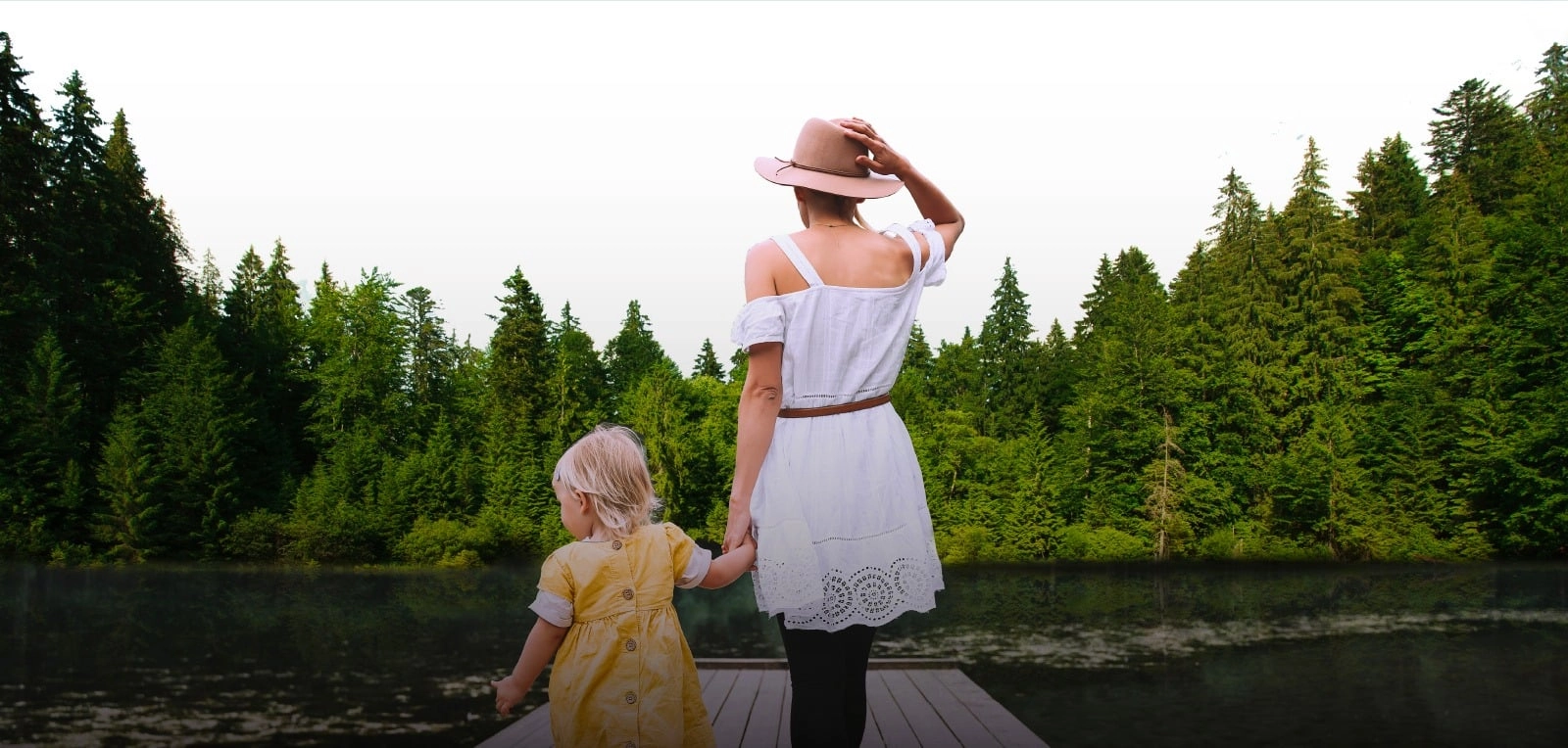 A woman and child holding hands on a dock beside a forest-lined lake.
